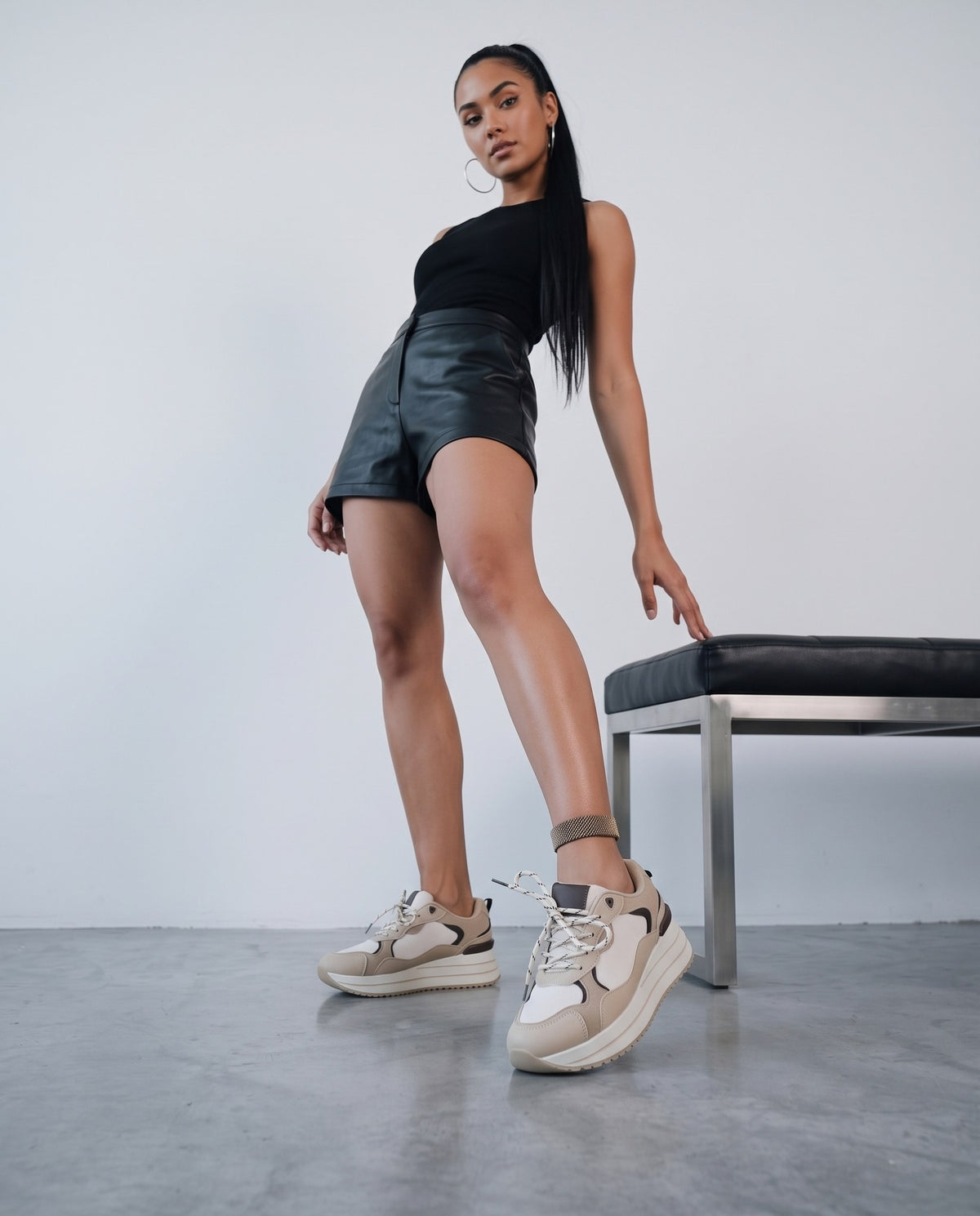 Modella in piedi con sneakers khaki/beige, panca metallica, outfit nero coerente con la serie.