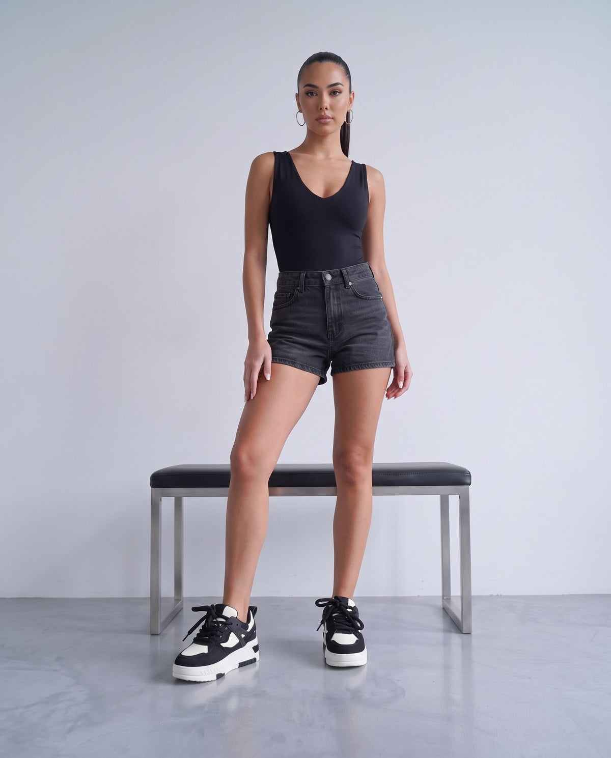 Modella in piedi con sneaker bicolore nero/bianco, body nero e shorts in denim scuro, panca in acciaio sullo sfondo