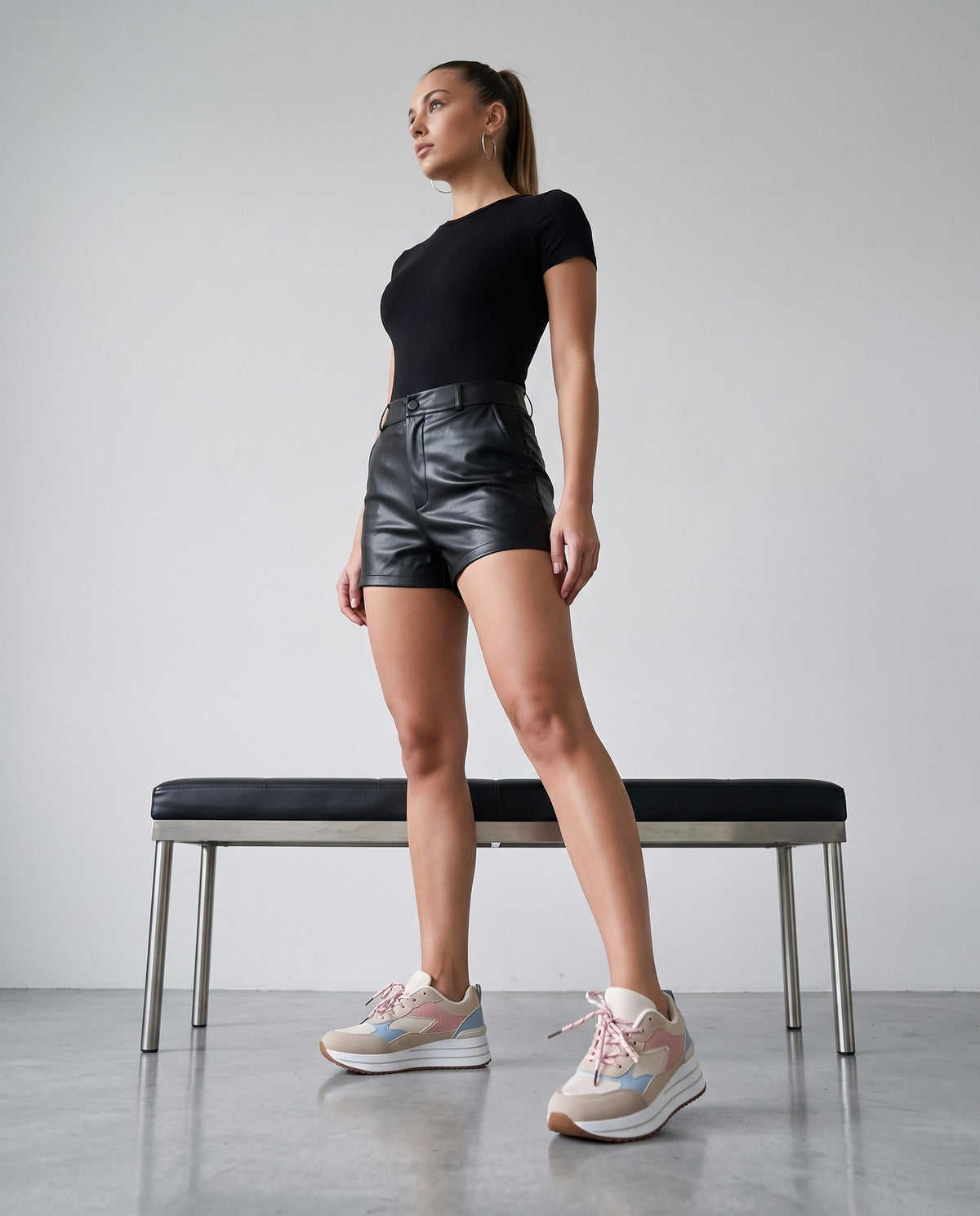 Modella in piedi con body nero, shorts neri e sneakers beige-rosa-azzurro, accanto a una panca nera.