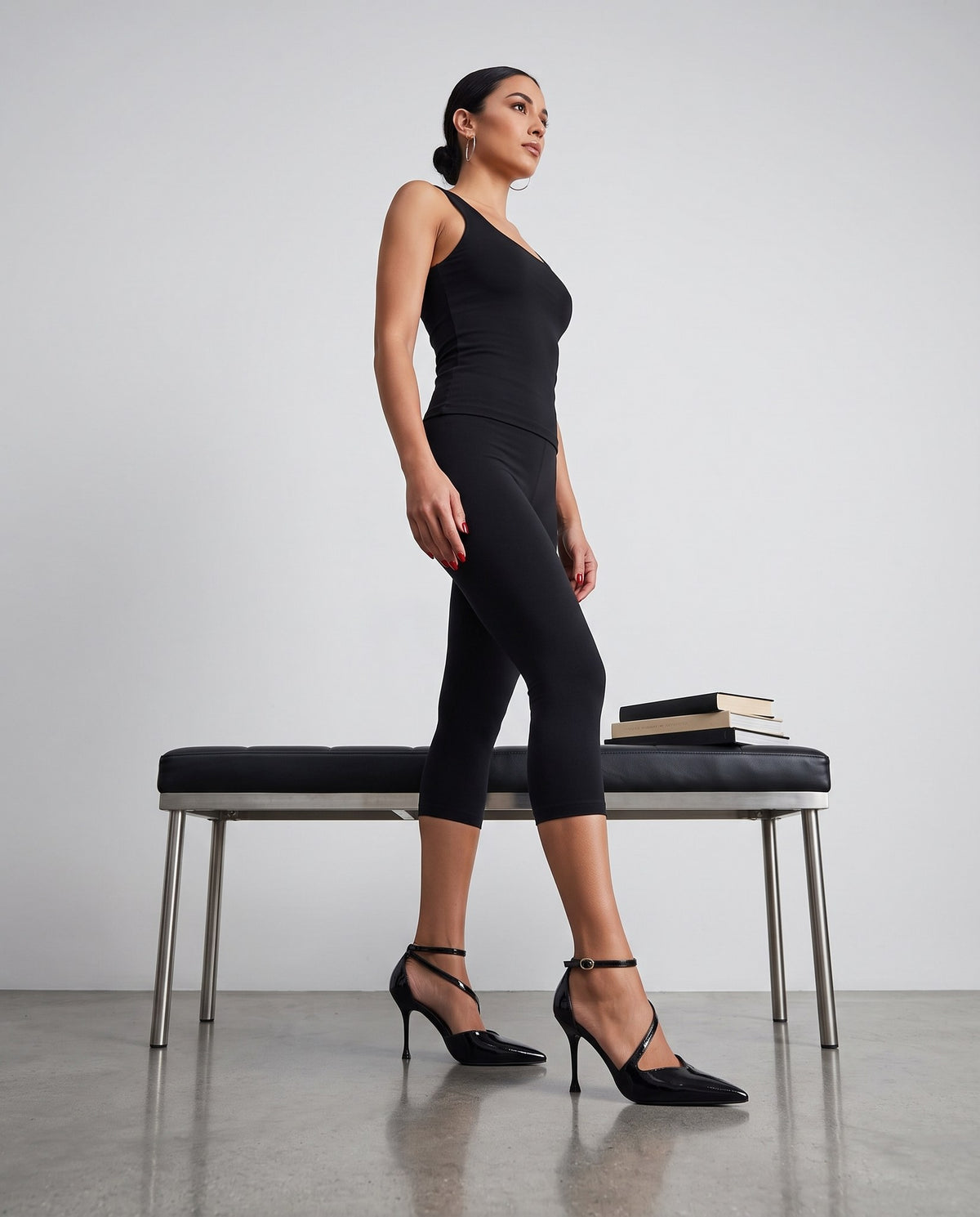 Modella in piedi di profilo con canotta e leggings neri, décolleté nere lucide con cinturino alla caviglia, panca in pelle sullo sfondo.