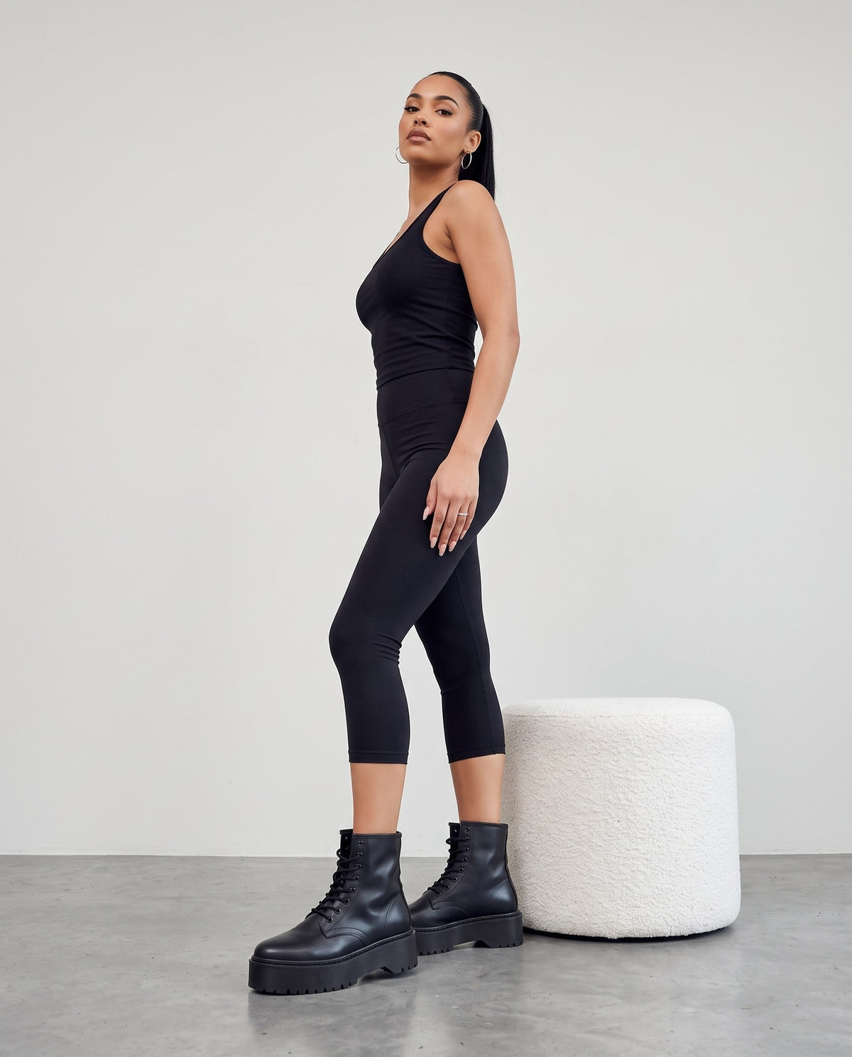 Modella di profilo con stivaletti neri platform, canotta e leggings neri, accanto a pouf bianco bouclé.