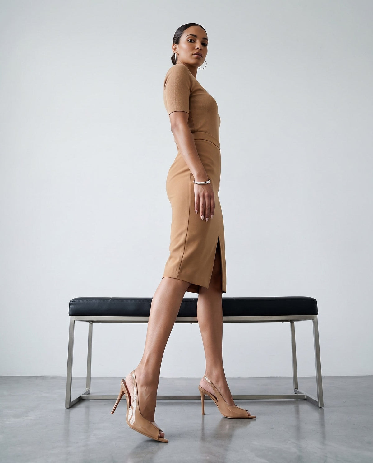 Modella di profilo con tubino cammello e décolleté slingback beige vicino a panca nera e cromata