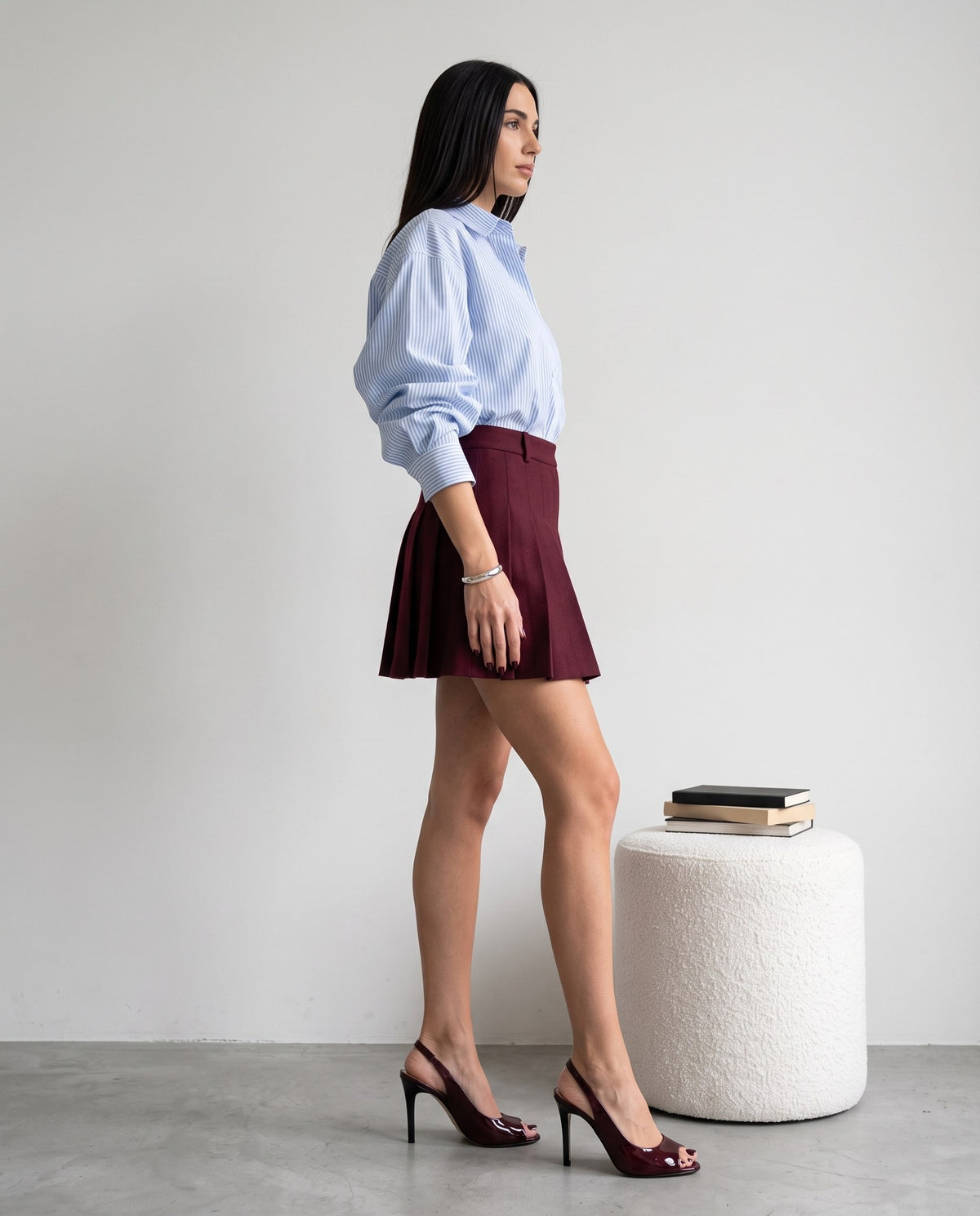 Modella di profilo con camicia a righe e minigonna bordeaux, sandali slingback bordeaux vernice, accanto a pouf bianco con libri