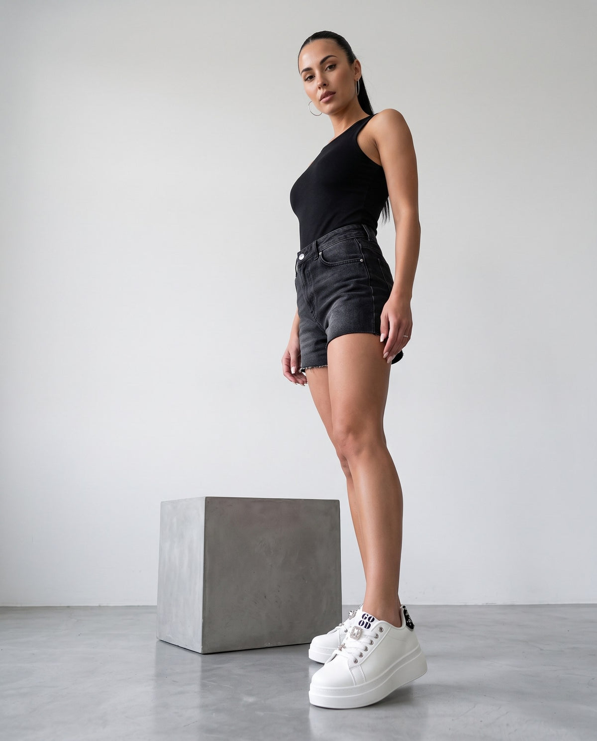 Modella in piedi con canotta nera, shorts di jeans neri e sneaker bianche platform, accanto a un blocco di cemento.