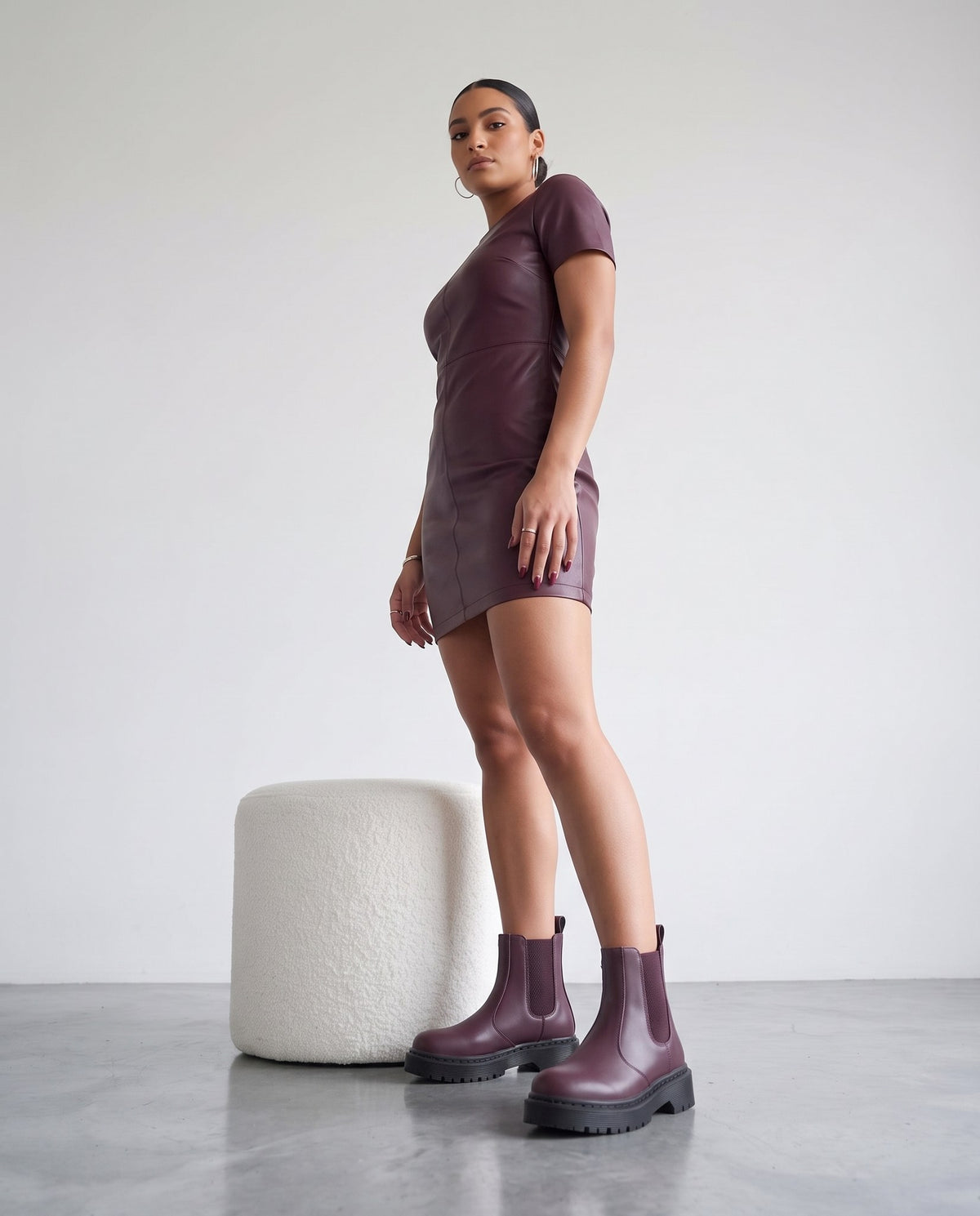 Modella in abito di pelle bordeaux e stivaletti chelsea bordeaux con suola nera, in piedi accanto a pouf bianco
