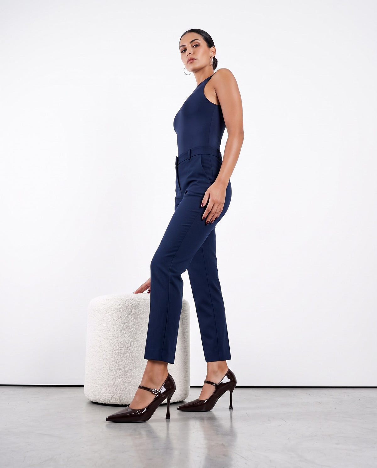 Modella in tuta blu notte indossa décolleté marrone scuro lucido con cinturino, accanto a pouf bianco