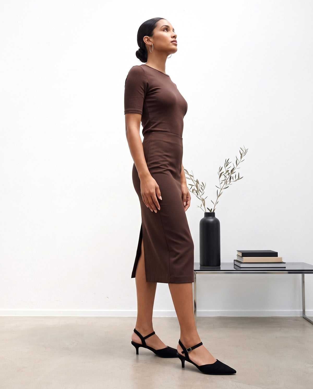 Modella di profilo con abito cioccolato e décolleté nere slingback con tacco basso, in studio con vaso e tavolino.