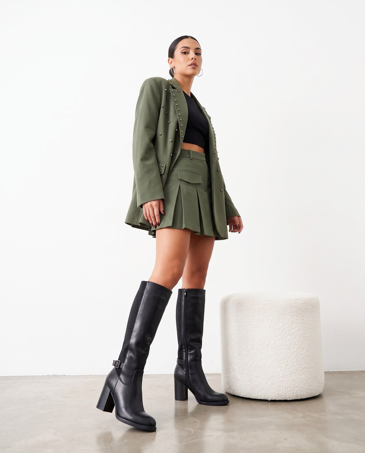 Modella in piedi con blazer verde militare, minigonna plissettata e stivali neri al ginocchio, accanto a pouf bianco.
