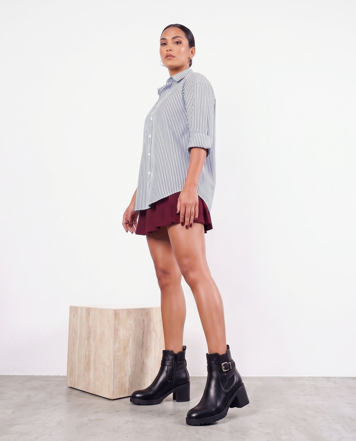 Modella di profilo con stivaletti neri con fibbia, camicia a righe e minigonna bordeaux, accanto a cubo in travertino