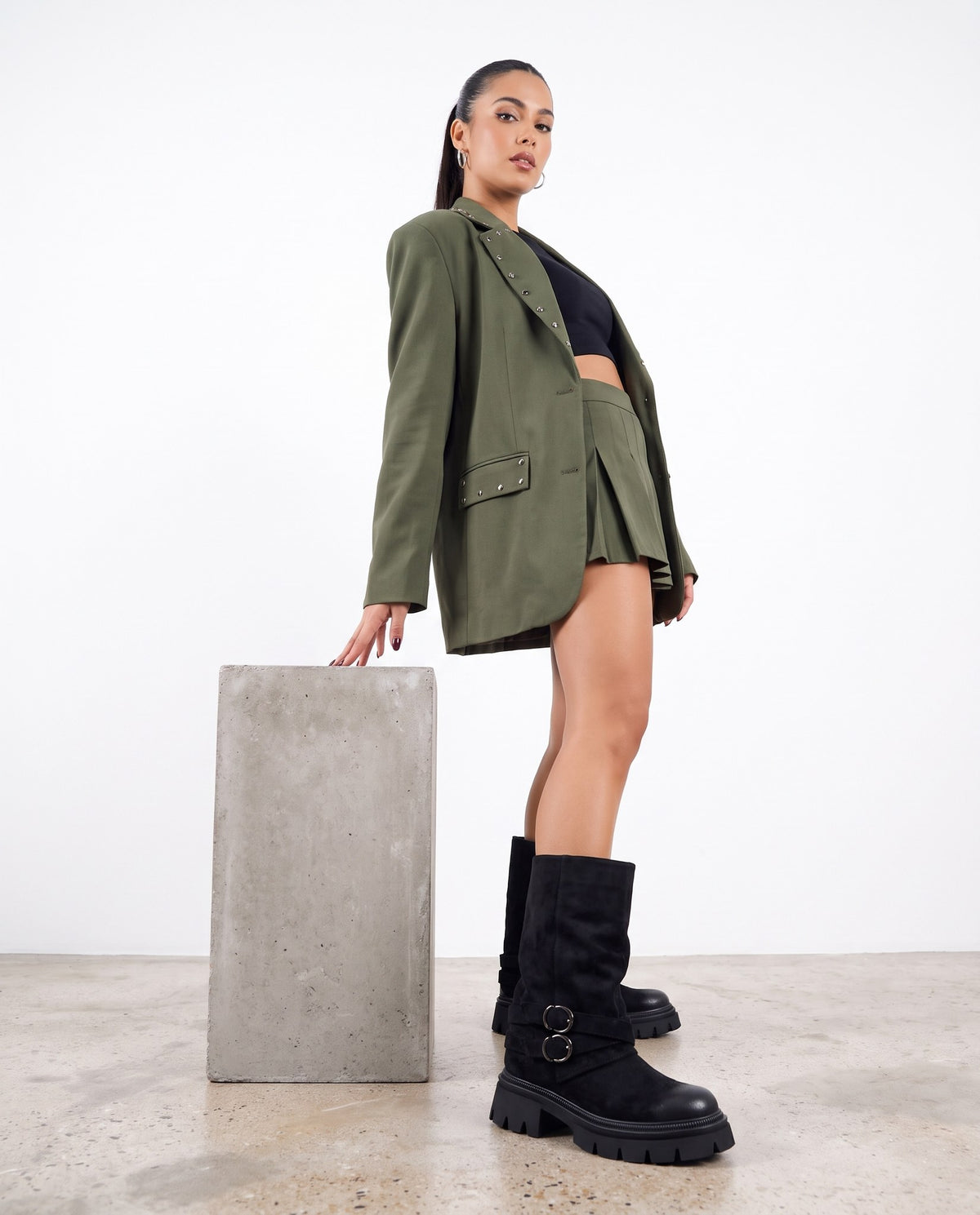 Modella in blazer e minigonna verde militare con stivali neri camoscio, accanto a blocco di cemento