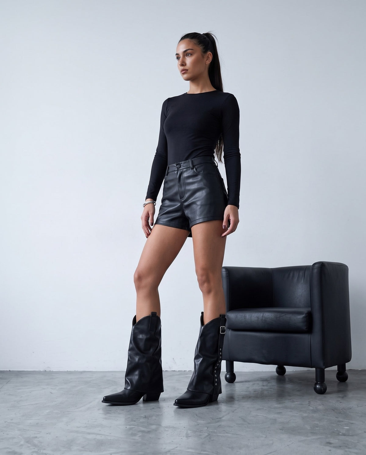 Modella in piedi con stivali neri cowboy con gambaletto, fibbia e borchie, accanto a poltrona nera.