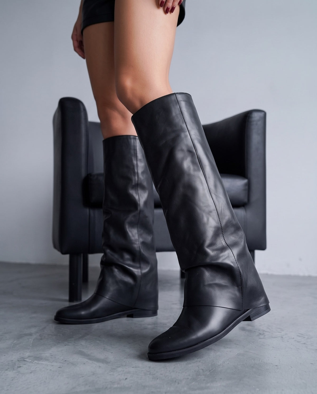 Dettaglio di due stivali alti in pelle nera con gambale morbido, su gambe femminili, sfondo poltrona nera sfocata.