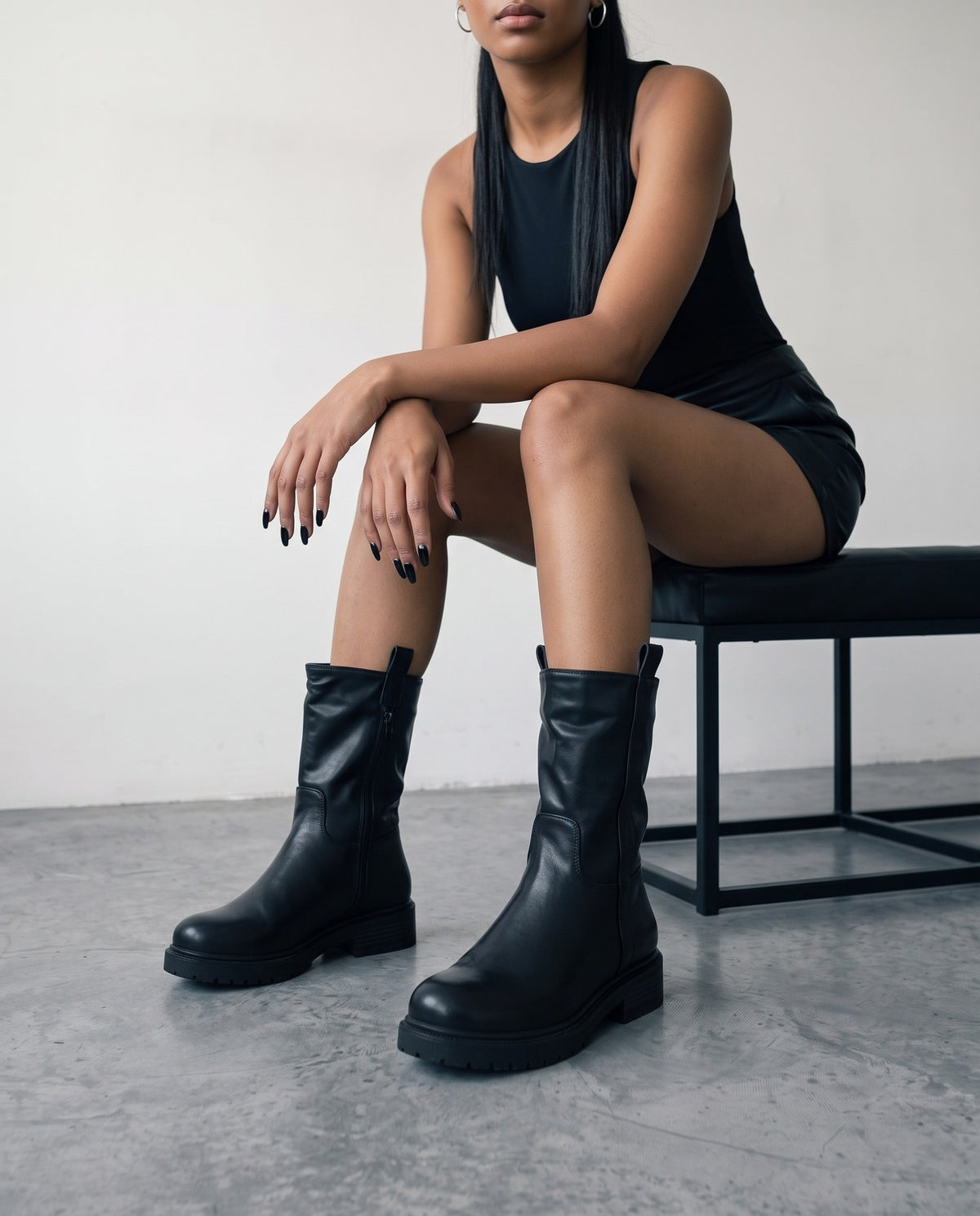 Modella seduta su panca nera con stivali biker neri in primo piano, gambe accavallate.