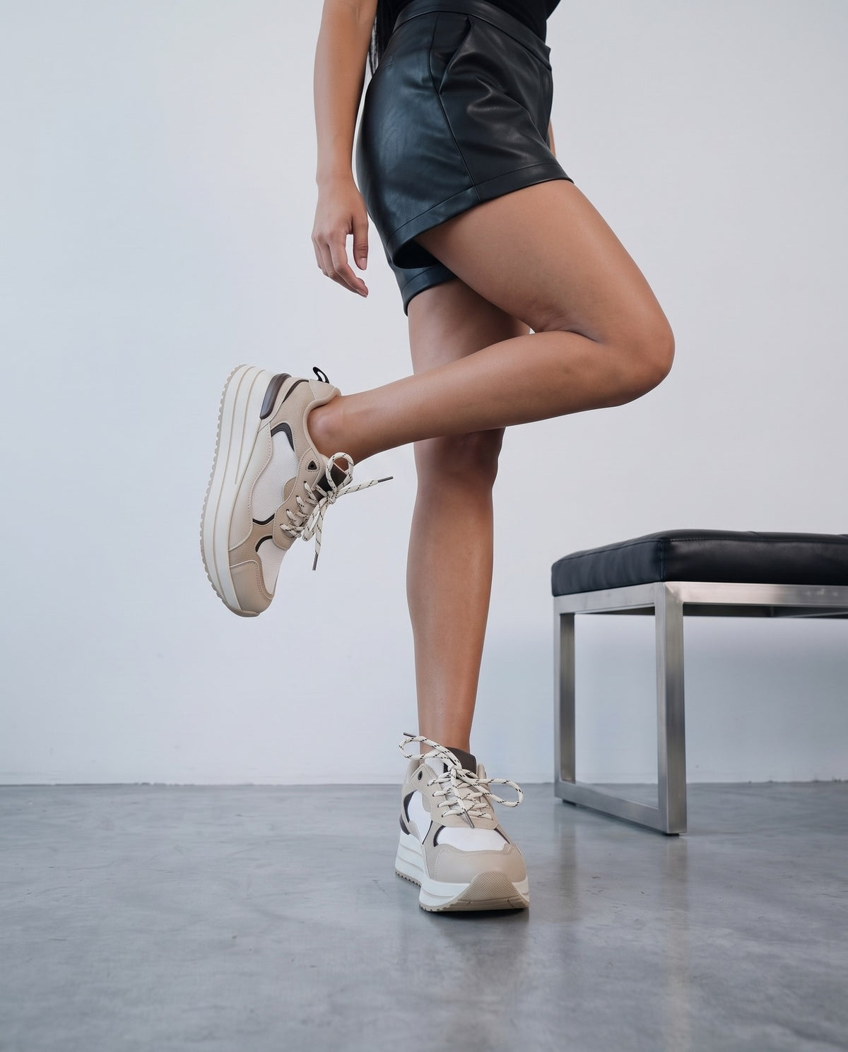 Modella in piedi con una gamba sollevata, sneaker khaki/beige platform bicolore, set con panca nera