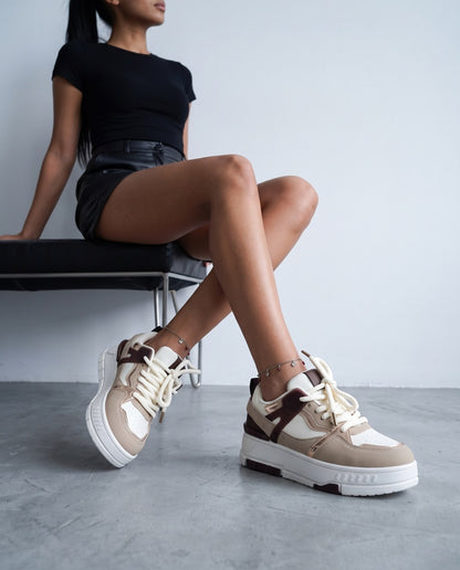 Modella seduta su panca nera, gambe accavallate, sneaker bicolore beige/bianco in primo piano