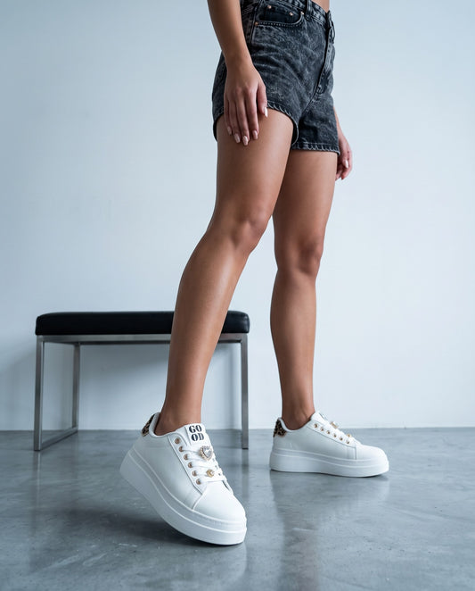 Gambe in piedi con sneakers bianche dettaglio leopardato, posa incrociata, arredo in secondo piano.