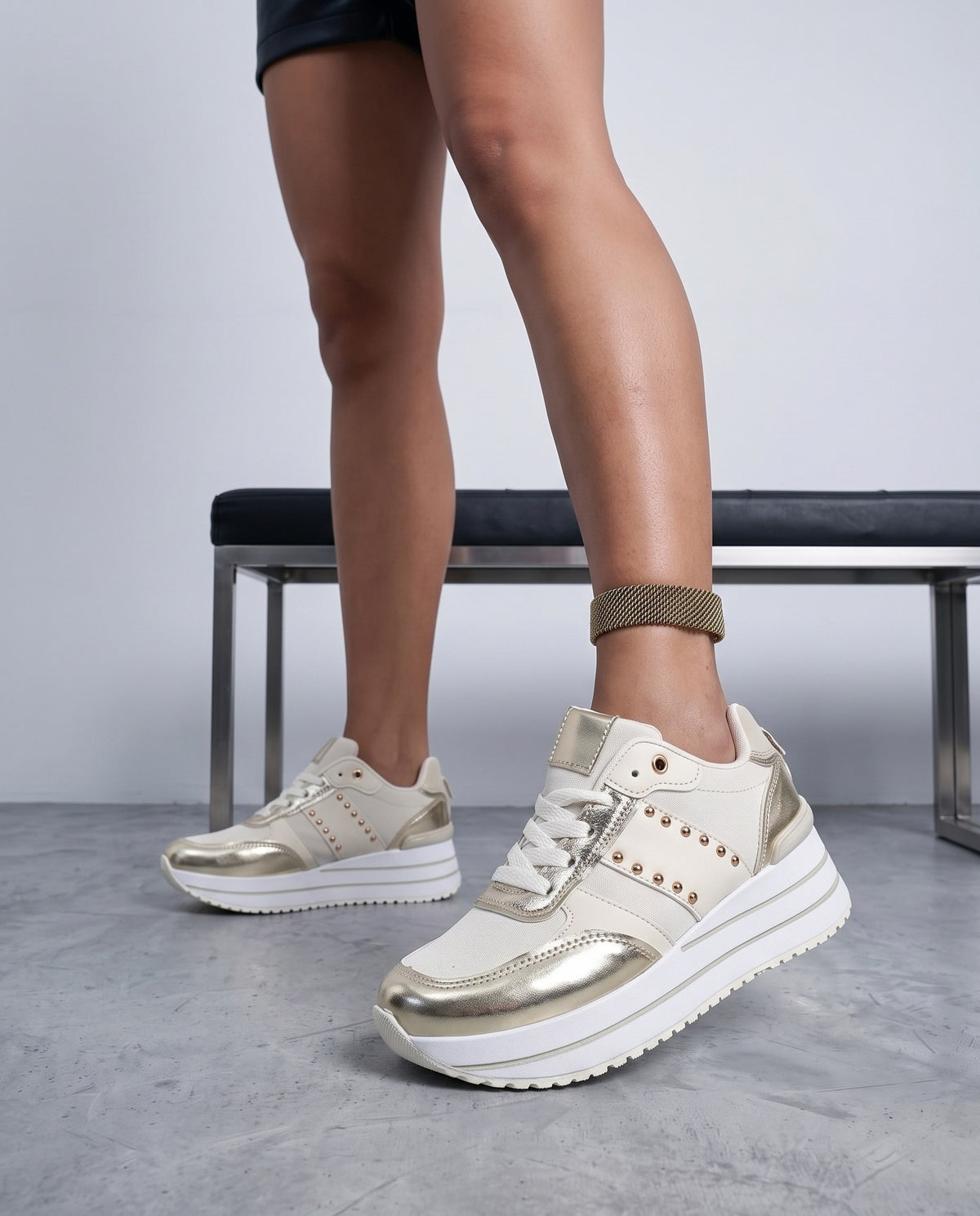 Primo piano di gambe con sneaker beige e oro, panca metallica sullo sfondo sfocato.