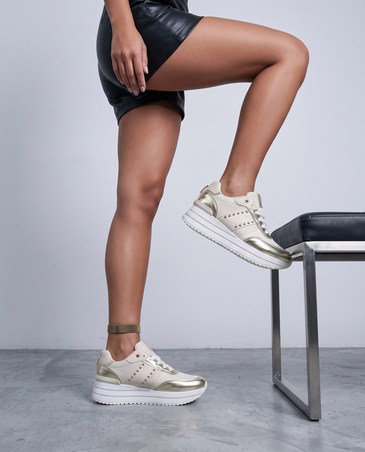 Donna in shorts di pelle nera con sneaker beige e oro, gamba piegata su panca, scatto dall'anca in giù.