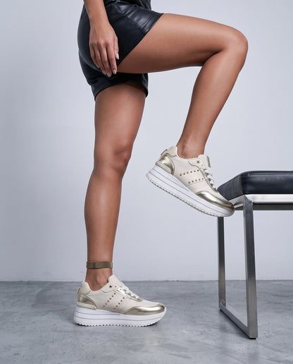 Donna in shorts di pelle nera con sneaker beige e oro, gamba piegata su panca, scatto dall'anca in giù.