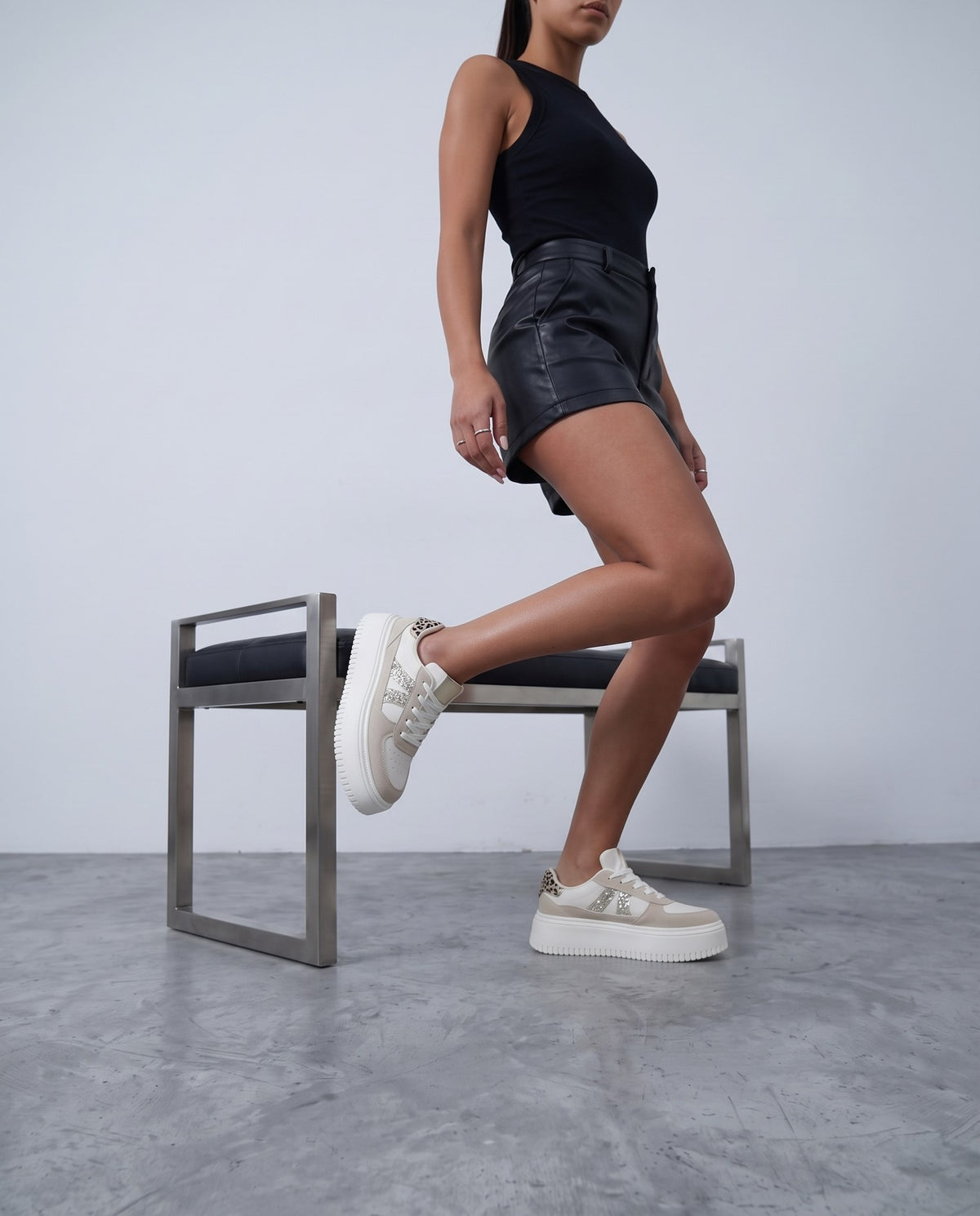 Modella in canotta nera e shorts in pelle, piede sollevato su sgabello, sneaker platform beige con glitter.