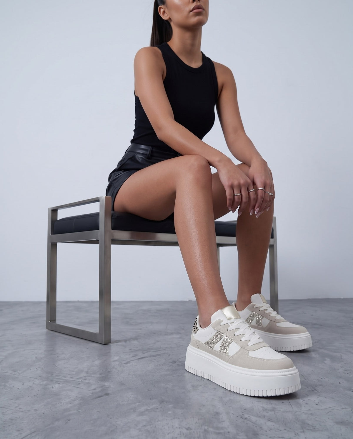 Modella seduta su sgabello, gambe incrociate alle caviglie, sneaker platform bianche e beige con glitter e tallone leopardato