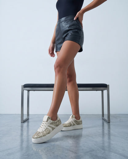 Gambe di modella con sneaker glitter oro/argento, suola platform bianca, accanto a panca in acciaio e pelle nera.