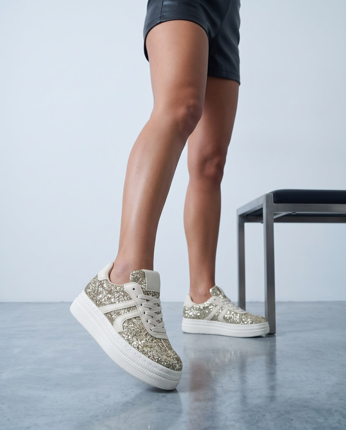 Primo piano di gambe con sneaker glitter oro e suola bianca platform, panca in metallo sullo sfondo sfocata.