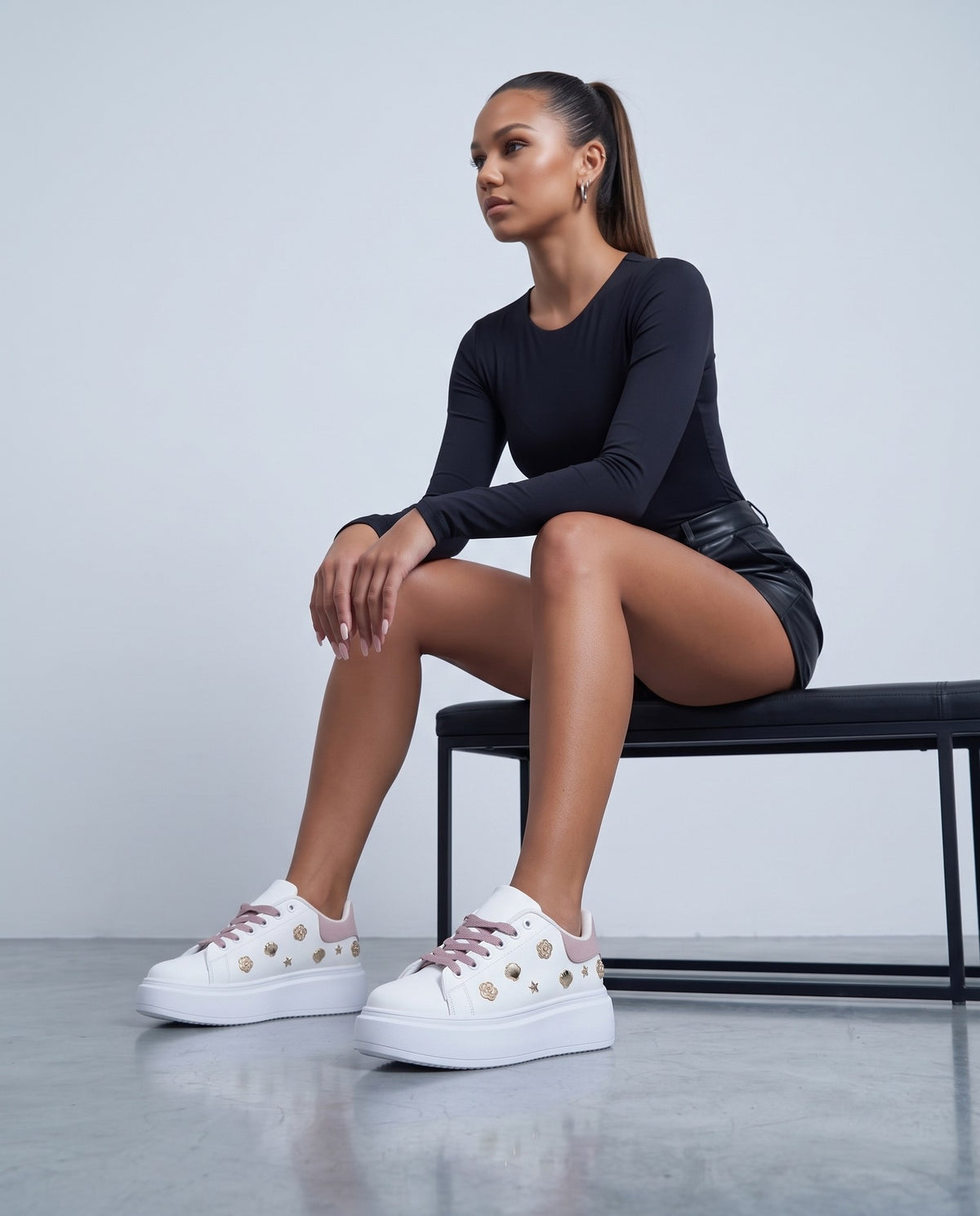 Modella seduta su panca nera, sneakers bianche con dettagli rosa e borchie dorate in primo piano