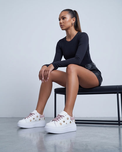 Modella seduta su panca nera, sneakers bianche con dettagli rosa e borchie dorate in primo piano
