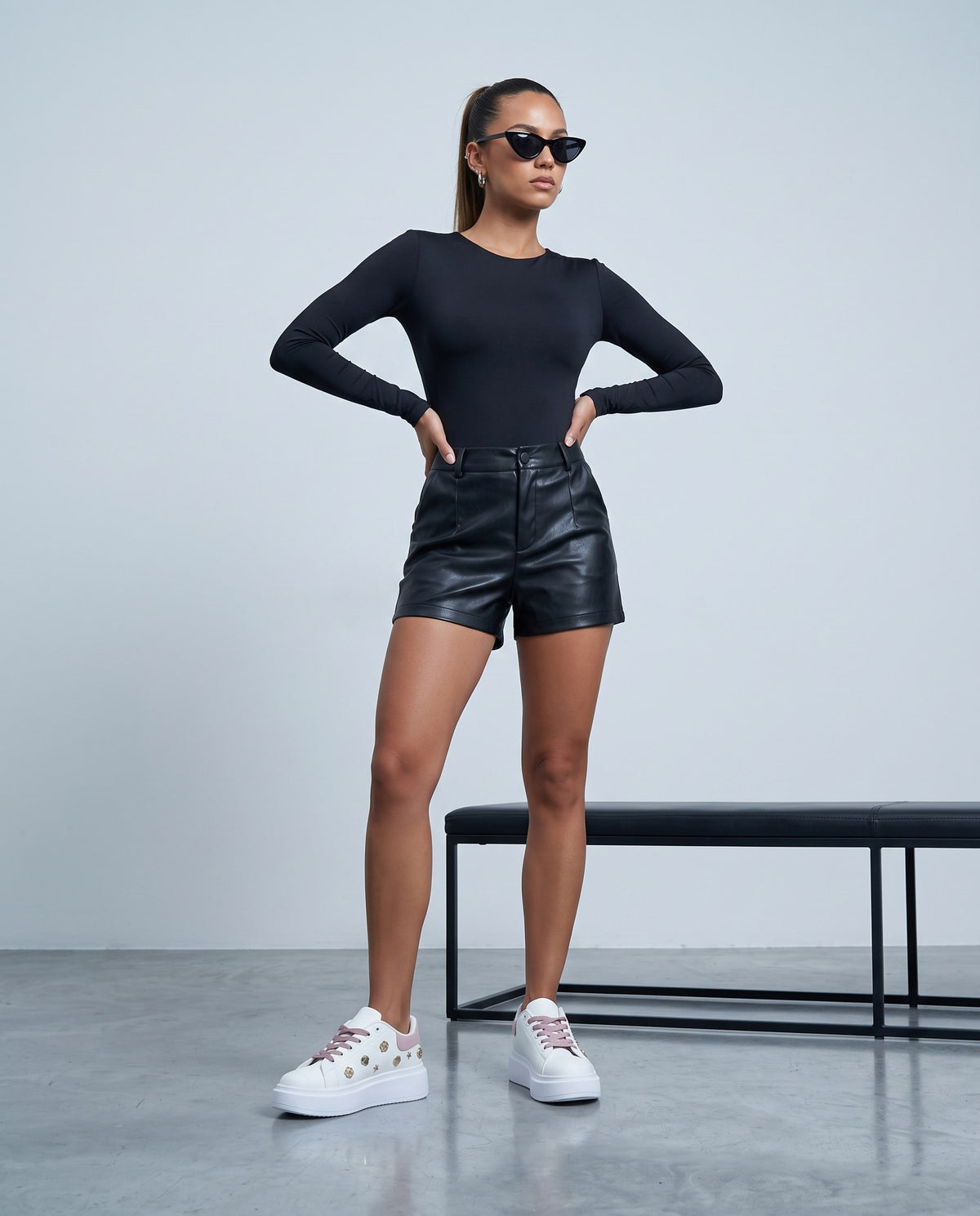 Modella in piedi con sneaker bianche e rosa con dettagli dorati, body nero e shorts neri.