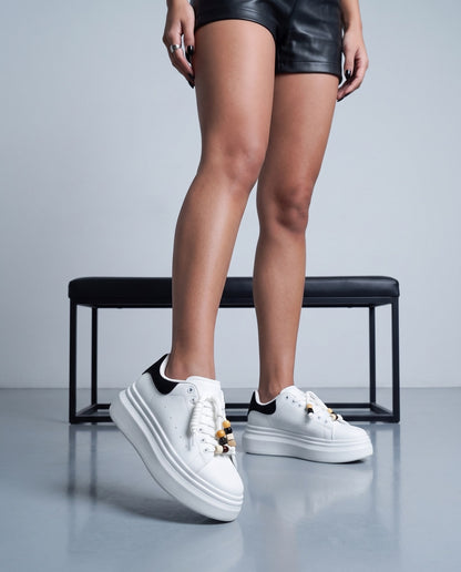 Dettaglio ravvicinato di gambe e sneaker bianche con tallone nero e perline sui lacci, panca nera sullo sfondo