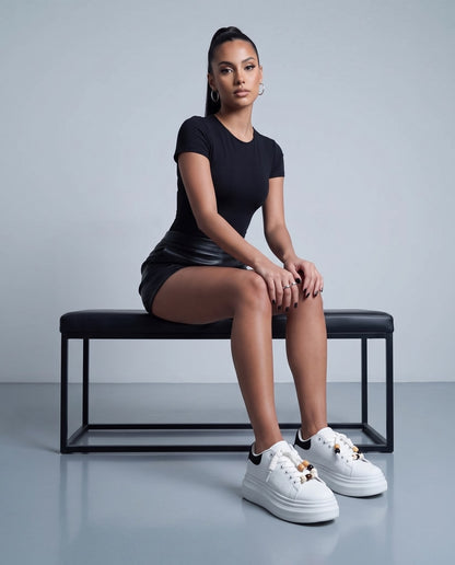 Modella seduta su panca indossa sneaker bianche platform con tallone nero e lacci con perline, body nero e mini shorts