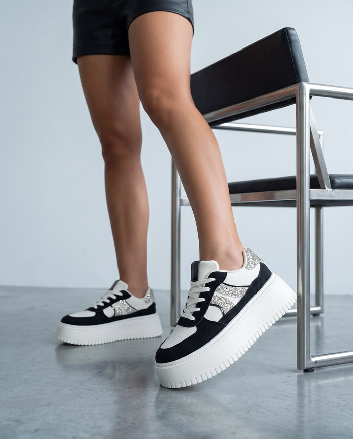 Dettaglio ravvicinato di gambe e sneaker bianche e nere con glitter, accanto a una sedia metallica.