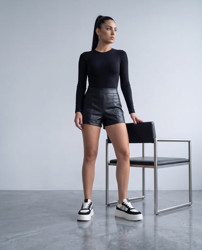 Modella in piedi con body nero e shorts in ecopelle, accanto a sedia, indossa sneaker platform bianche e nere glitterate