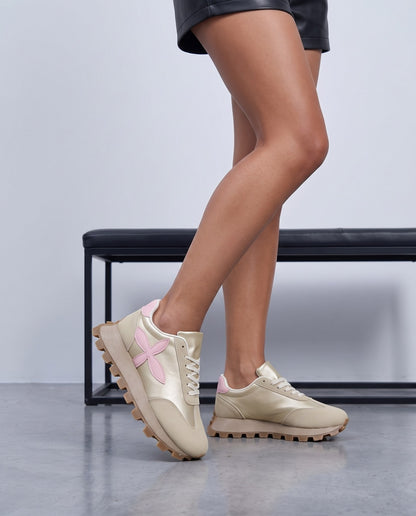 Dettaglio ravvicinato di gambe incrociate con sneakers beige dorate e fiore rosa, sfondo panca nera sfocata