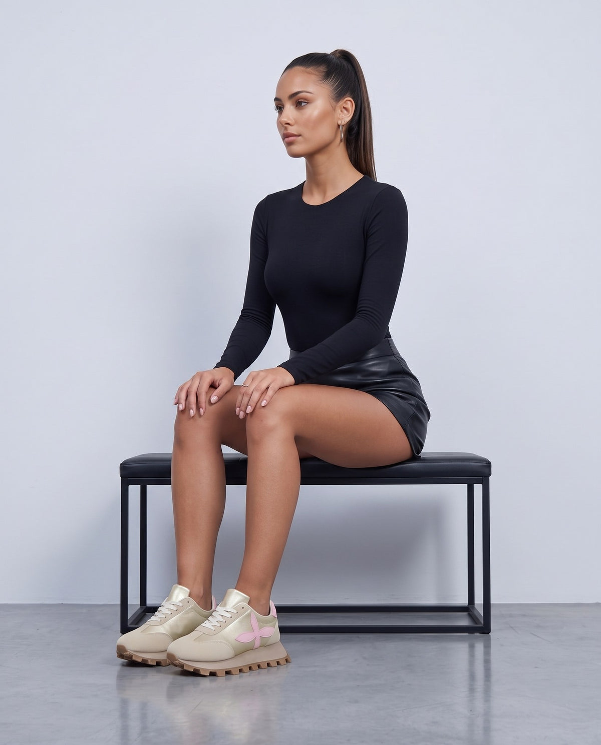 Modella seduta su panca nera, sneakers beige con fiore rosa, gambe accavallate, sfondo grigio chiaro