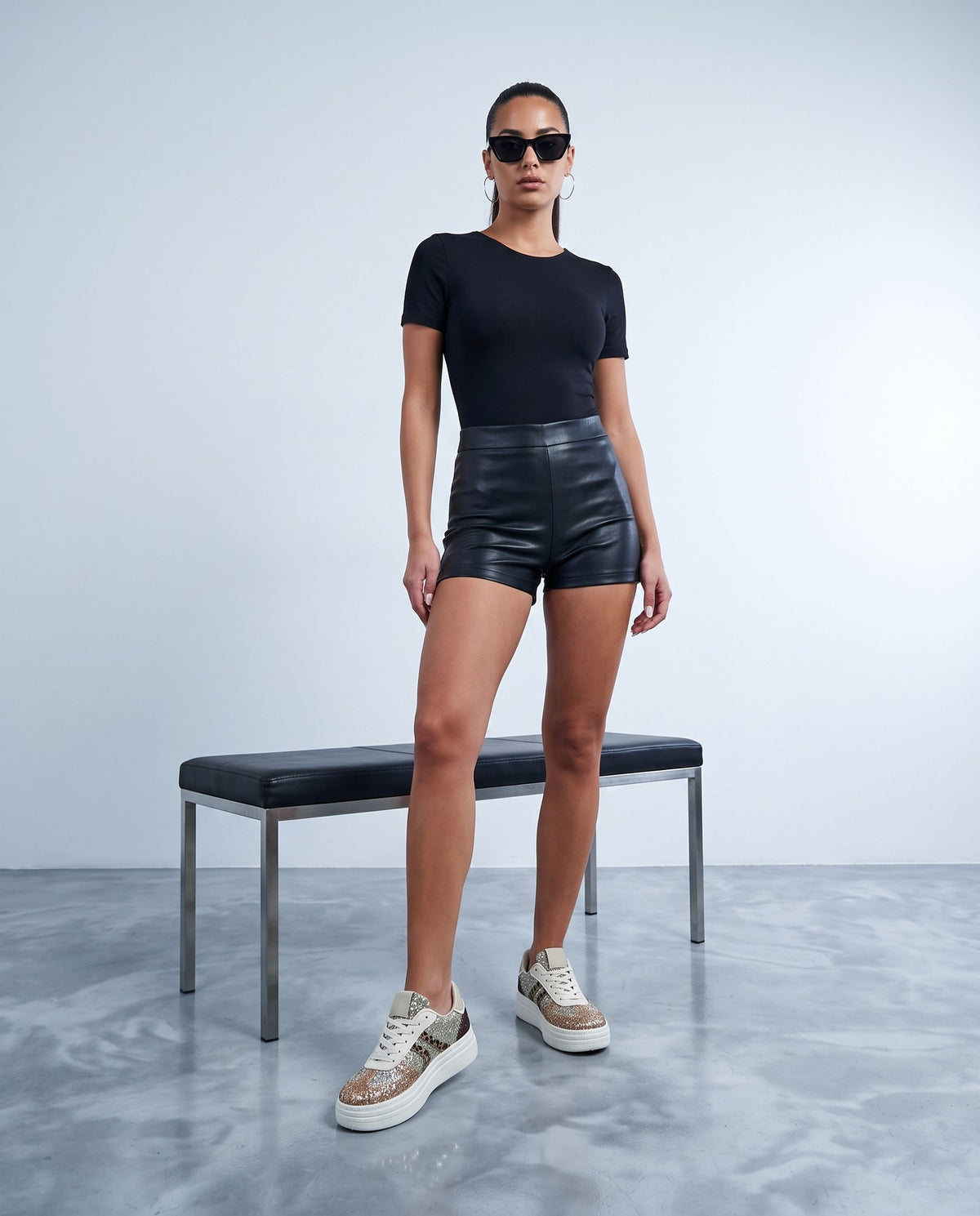 Modella in piedi con shorts e body neri, sneaker glitter multicolor con dettagli leopardati, panca in acciaio e pelle sullo sfondo