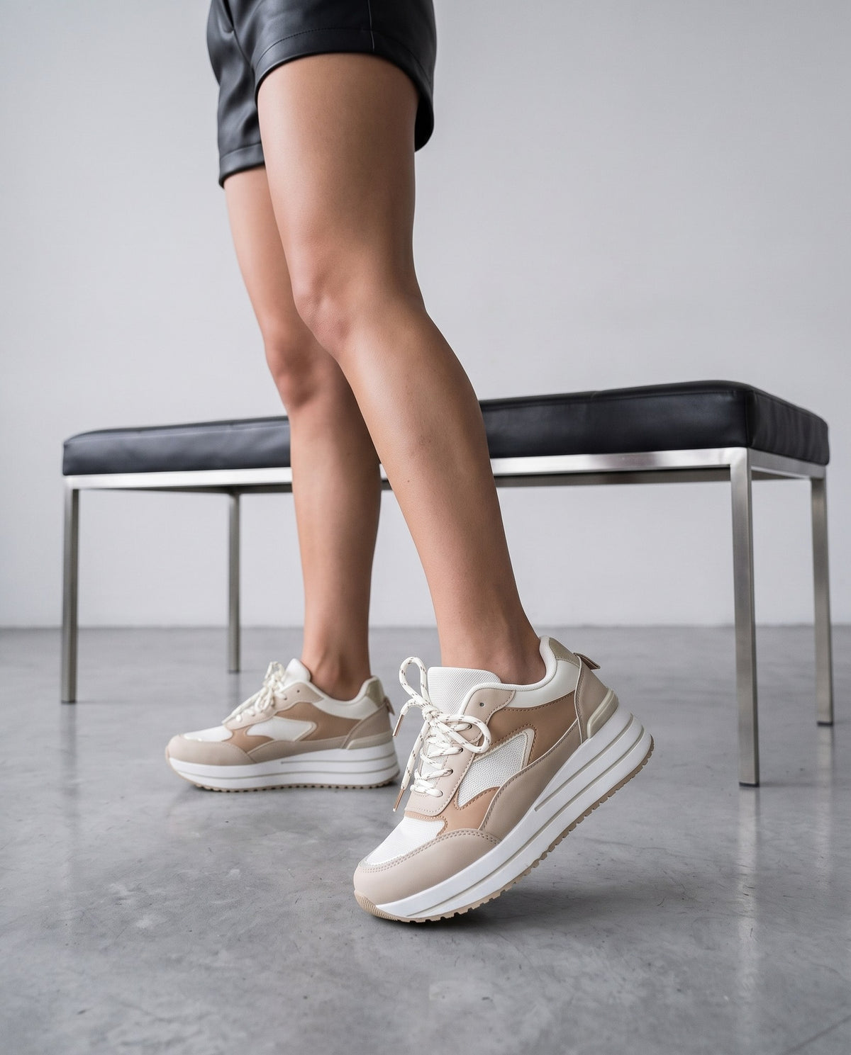 Gambe femminili con sneakers beige/bianche a pannelli, in posizione di passo, accanto a una panca nera, su pavimento in cemento chiaro.