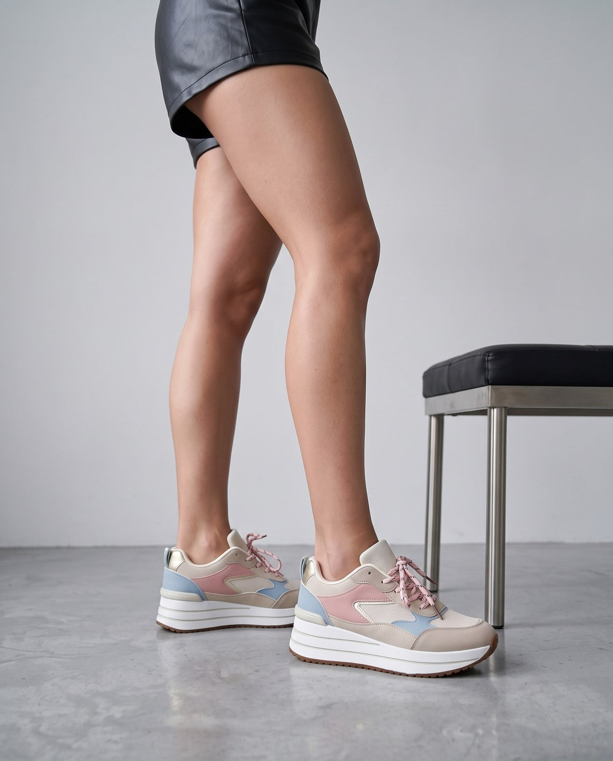 Gambe di modella in piedi con sneakers beige rosa e azzurro, panca nera accanto, pavimento in cemento