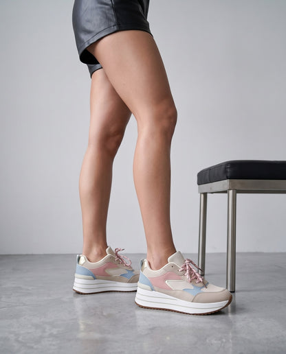 Gambe di modella in piedi con sneakers beige rosa e azzurro, panca nera accanto, pavimento in cemento