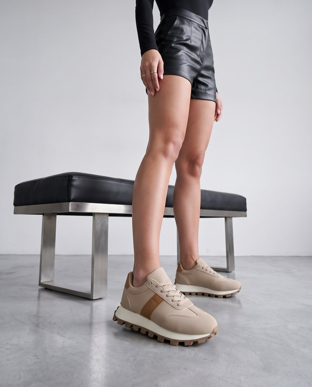 Gambe di modella in piedi con sneaker beige e cammello, accanto a panca nera in pelle e metallo.