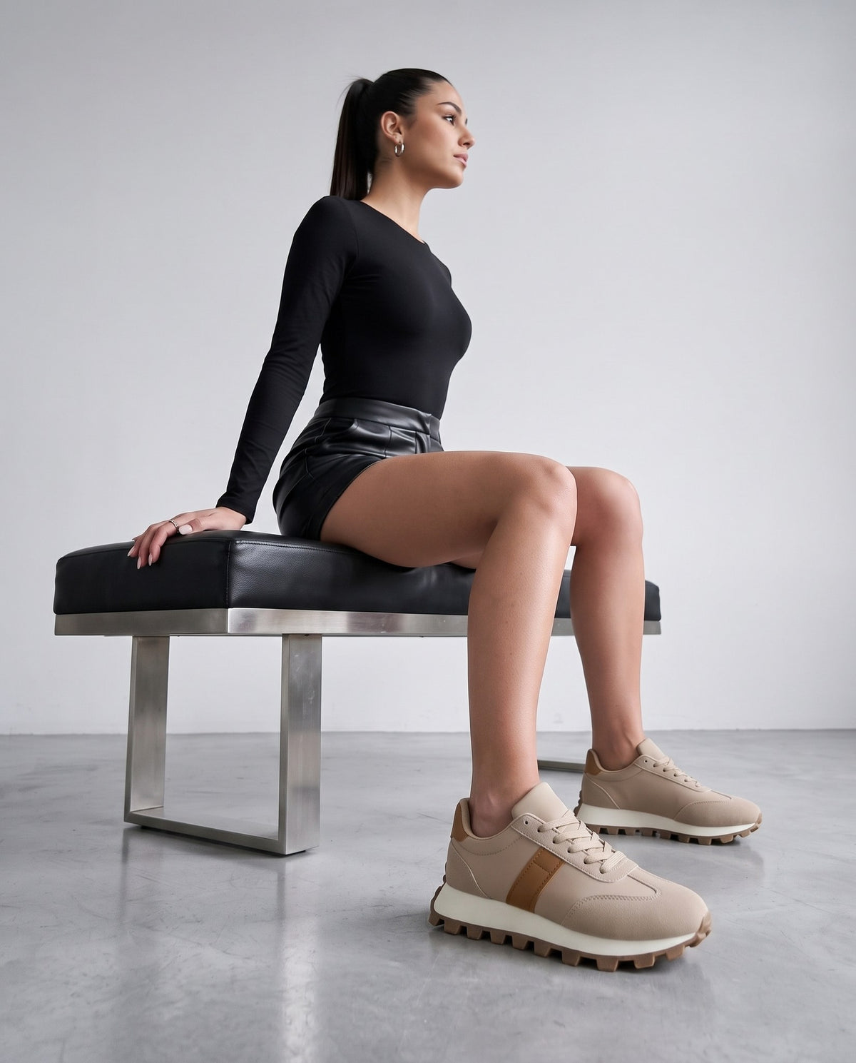 Modella seduta su panca nera, gambe incrociate, sneakers beige con dettagli cammello in primo piano.
