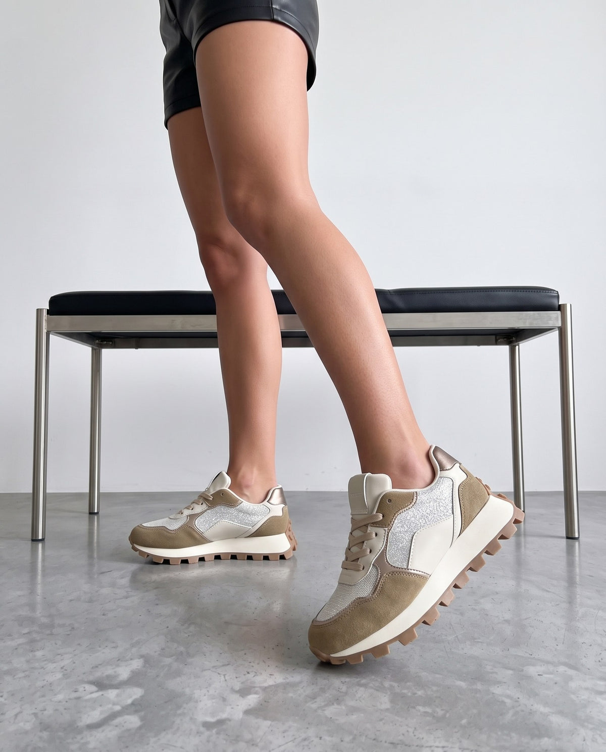 Gambe femminili con sneakers khaki glitter accanto a una panca in pelle nera su pavimento in cemento