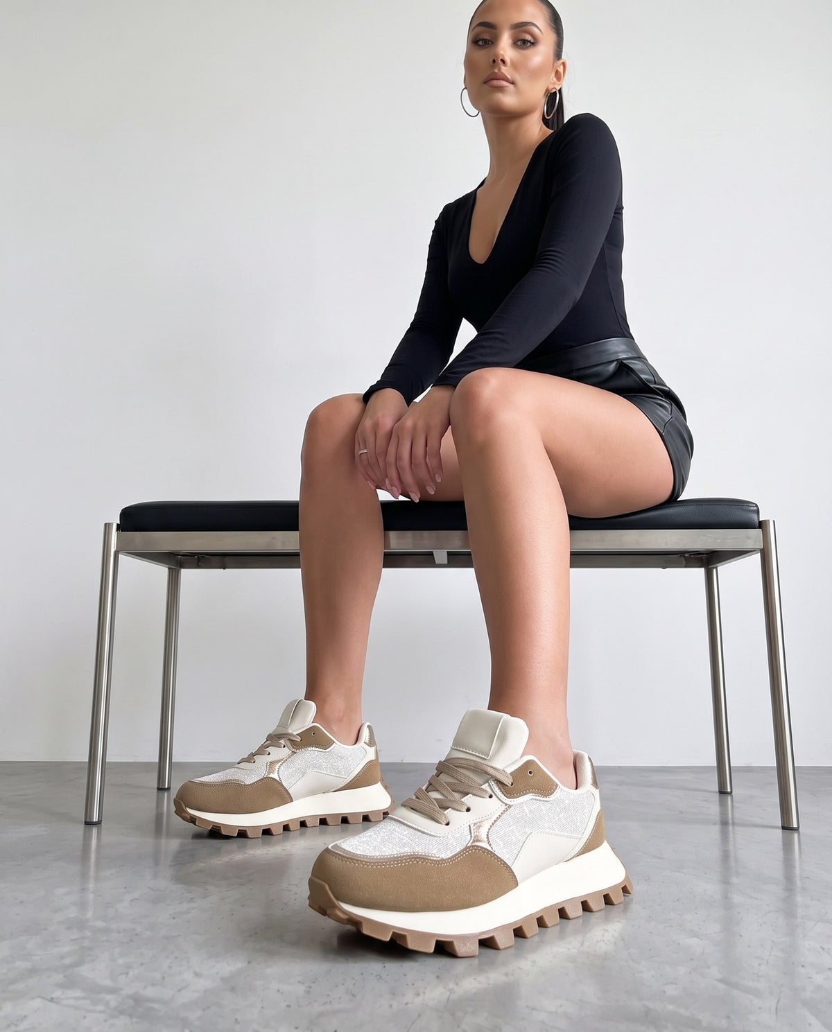 Modella seduta su panca, scarpe sneaker bianche e cachi in primo piano, gambe accavallate.