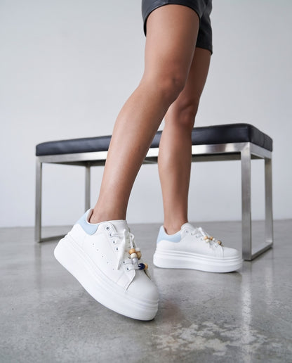 Dettaglio ravvicinato di gambe con sneaker bianche e azzurre con perline sui lacci, panca in sottofondo.