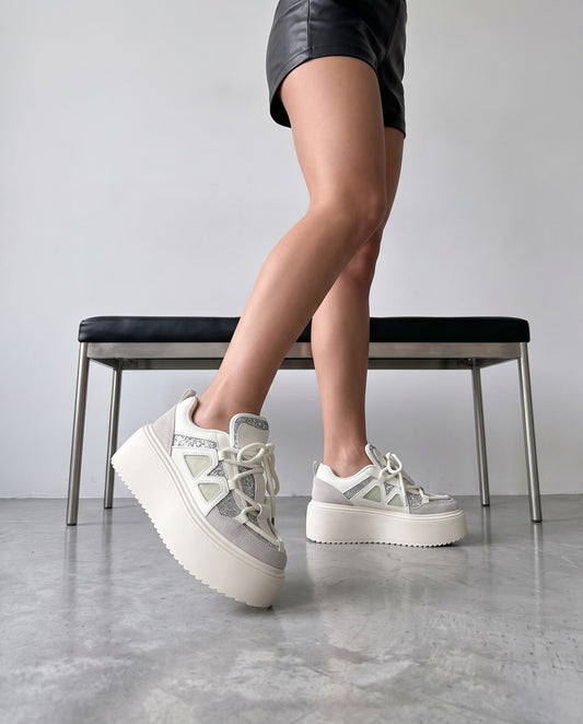 Gambe in piedi, sneaker bianca con glitter argento e inserti verdi, panca in acciaio e pelle nera sullo sfondo.