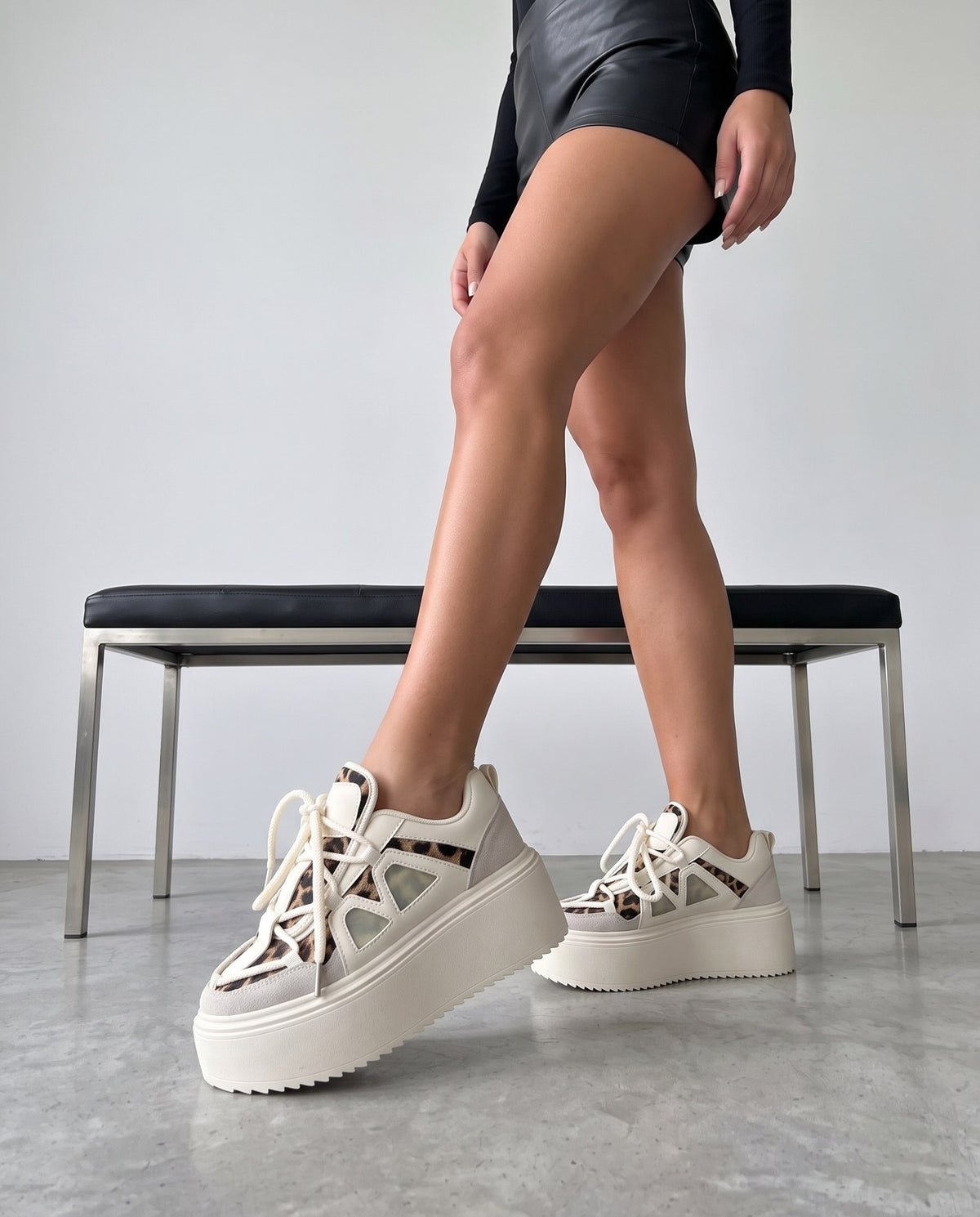 Gambe della modella con sneaker platform bianche e maculate, panca in acciaio e pelle nera sullo sfondo