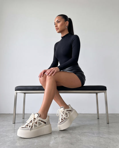 Modella seduta su panca nera, scarpe platform leopardate e bianche in primo piano, gambe accavallate.