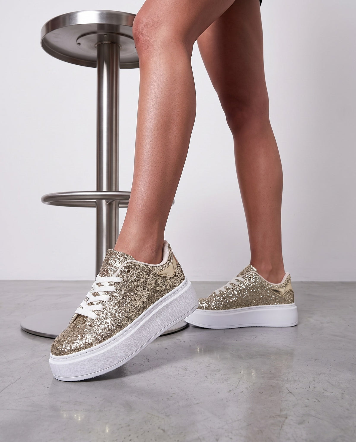 Dettaglio ravvicinato di due sneaker glitter oro con suola bianca, gambe nude, pavimento in cemento, sgabello in acciaio.