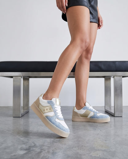 Gambe di modella in piedi che indossa sneakers bianche e denim con dettagli dorati, accanto a panca nera e metallo